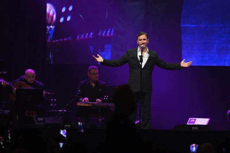 Kadim Al Sahir with open arms under purple stage light, his musicians beside him
