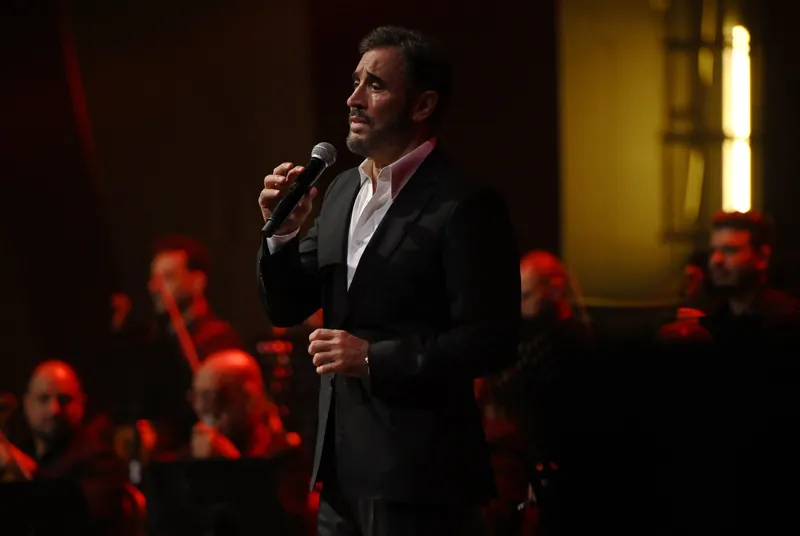 Kadim Al Sahir singing in warm light, the orchestra behind him